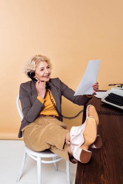 mujer mayor con estilo con las piernas en la mesa hablando en el teléfono giratorio y mirando el papel
  - Foto, Imagen