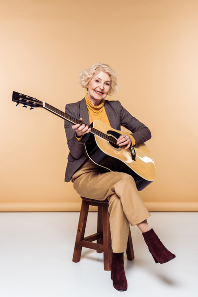 lächelnde, stylische Seniorin auf Stuhl sitzend und auf Akustikgitarre spielend  - Foto, Bild