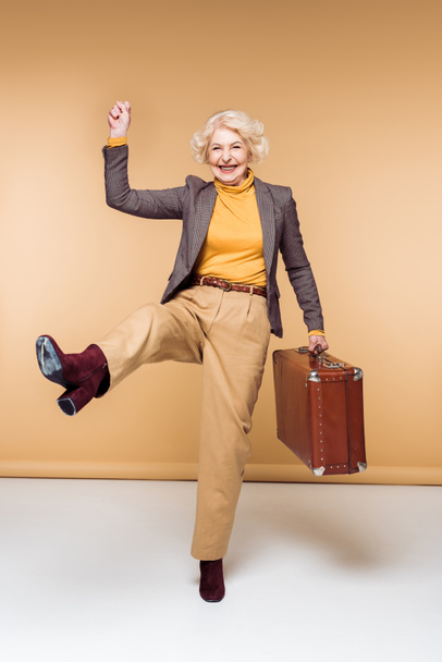 ενθουσιασμένος κομψό ανώτερος γυναικείο ταξιδιώτη με βραχίονα έθεσε κρατώντας vintage βαλίτσα  - Φωτογραφία, εικόνα