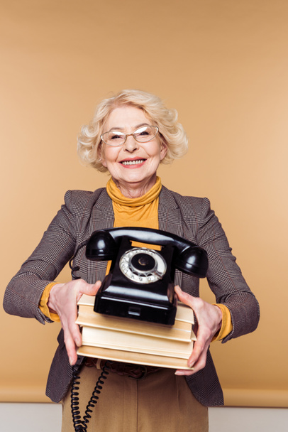 stylish senior woman in eyeglasses holding rotary phone and stack of books  - Zdjęcie, obraz