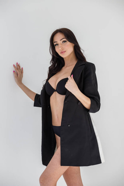 Studio portrait of young brunette girl wearing sexy black lingerie and black coat standing on white wall background - Fotoğraf, Görsel
