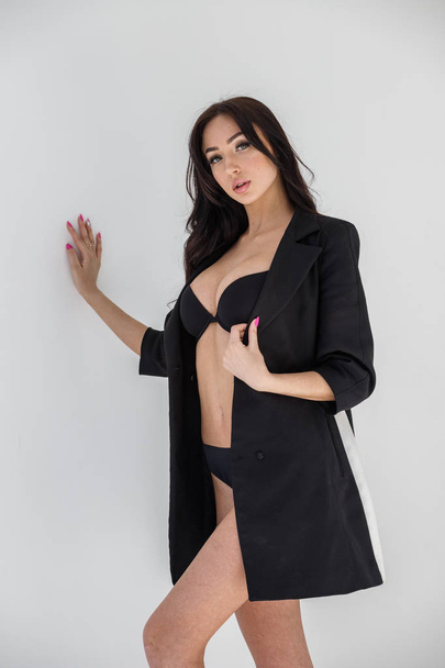 Studio portrait of young brunette girl wearing sexy black lingerie and black coat standing on white wall background - Fotó, kép