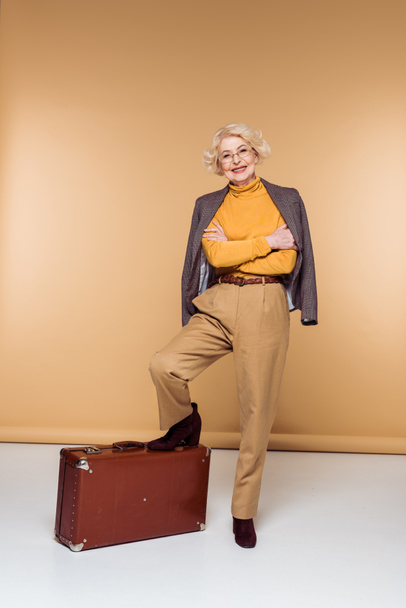 fashionable senior female tourist with jacket on shoulder holding leg on vintage suitcase  - Fotoğraf, Görsel
