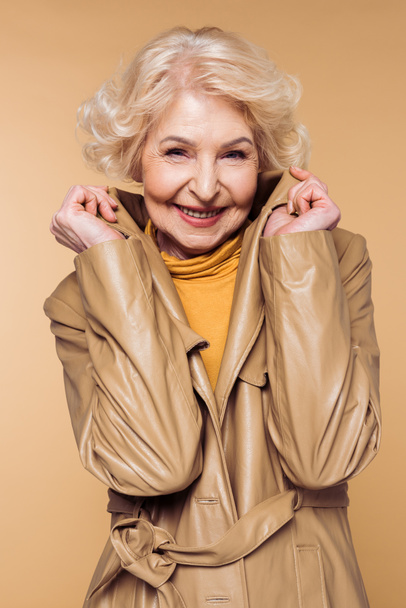 smiling stylish senior woman posing and holding collar of trench coat  - Foto, afbeelding