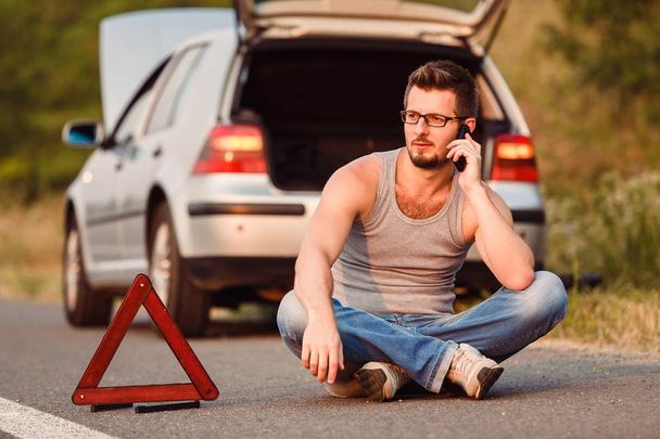 A young man sits on the asphalt, near his broken car and call technical support. Damage on the road - 写真・画像