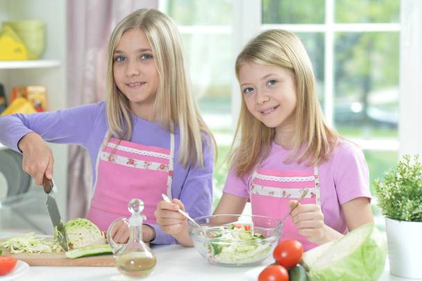Kaksi tyttöä esiliinoissa valmistelee tuoretta salaattia keittiön pöydällä
   - Valokuva, kuva