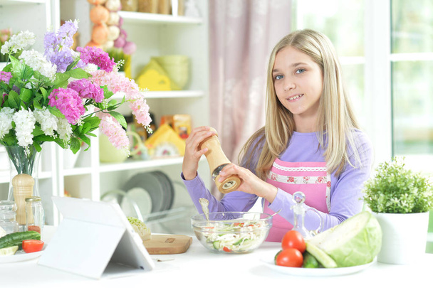Linda chica preparando ensalada fresca en la mesa de la cocina con la tableta en casa
 - Foto, Imagen