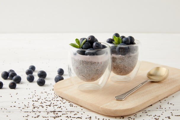 close up view of chia puddings with fresh blueberries and mint on wooden cutting board on white tabletop - Фото, зображення