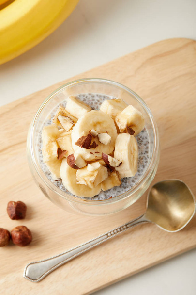 Blick von oben auf leckeren Chiasamen-Pudding mit Bananenstücken und Haselnüssen auf weißer Tischplatte - Foto, Bild