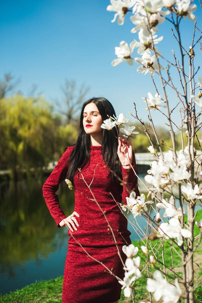 красивая юная брюнетка с длинными волосами в парке весной весной среди цветущих деревьев в красном платье в руках держит цветы магнолии
 - Фото, изображение