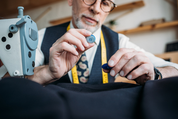 cropped image of handsome tailor choosing buttons at sewing workshop - Photo, Image