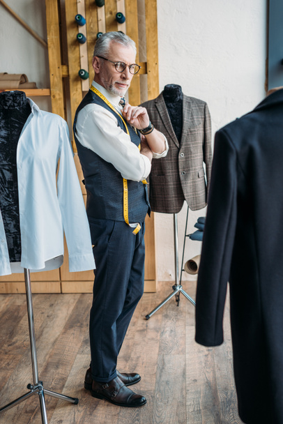 guapo sastre de pelo gris mirando chaquetas en maniquíes en taller de costura
 - Foto, Imagen