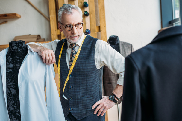 sastre guapo apoyado en el maniquí en el taller de costura y mirando chaqueta
 - Foto, Imagen