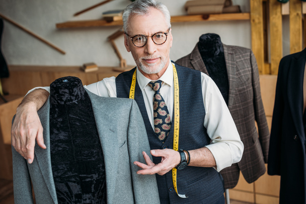 sastre guapo mostrando chaqueta en maniquí en taller de costura
 - Foto, Imagen