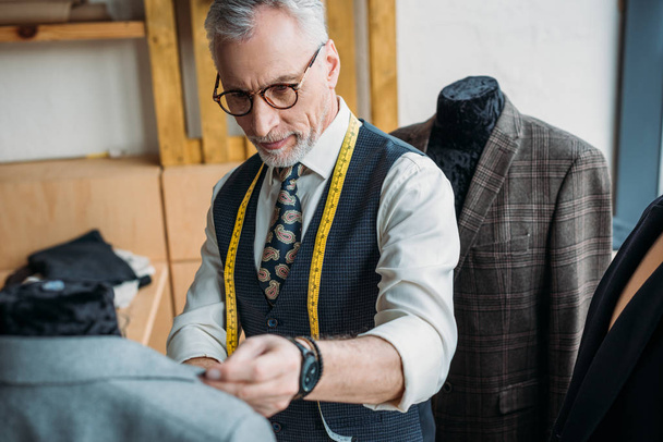 guapo sastre maduro con cinta métrica mirando chaqueta en maniquí en taller de costura
 - Foto, Imagen