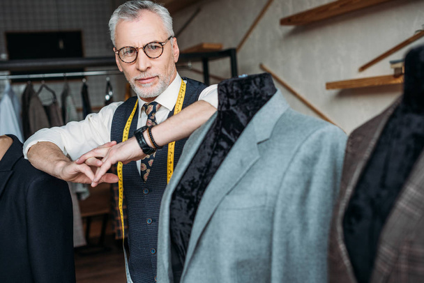 elegante sastre maduro con cinta métrica apoyada en maniquí con chaqueta en el taller de costura
 - Foto, Imagen
