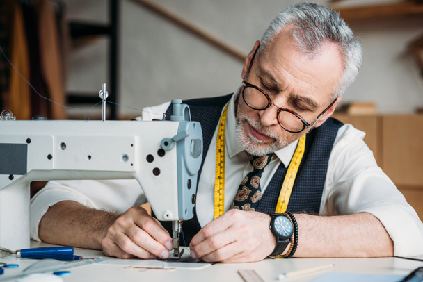 focused mature tailor working with sewing machine at workshop - 写真・画像