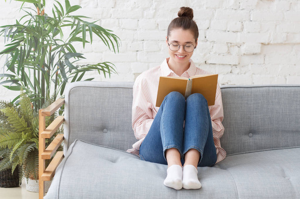 Indoor portrait of young beautiful European girl pictured on grey sofa in cozy room or studio with eyeglasses and casual clothes on, reading a book or looking through pages of copybook in hard cover - Valokuva, kuva