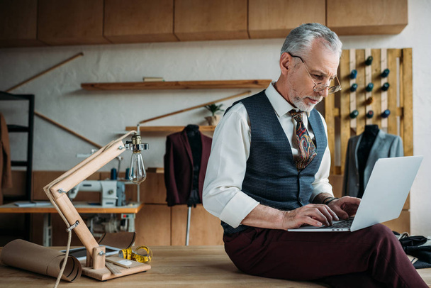 tailleur mature concentré travaillant avec ordinateur portable tout en étant assis sur la table à l'atelier de couture
 - Photo, image