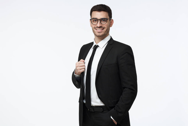 Half-length portrait of handsome Caucasian guy dressed in black suit, white shirt and tie isolated on gray background looking attentively through glasses and smiling positively and confidently - Zdjęcie, obraz