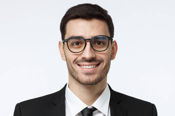 Closeup headshot of young handsome European businessman in glasses pictured isolated on gray background wearing black formal suit, white shirt and tie, looking confident and smiling friendly - Foto, Bild