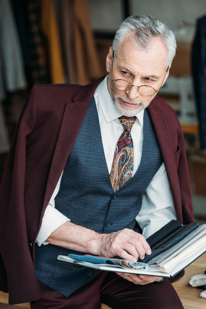 縫製工場でテーブルに座っている布タイプのカタログでスタイリッシュな成熟したテーラー - 写真・画像