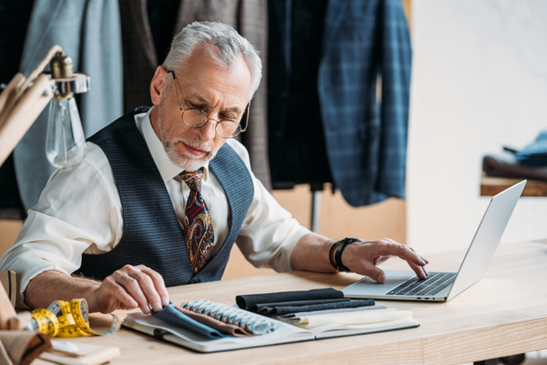 handsome mature tailor working with laptop and cloth samples at sewing workshop - 写真・画像