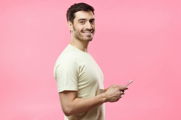 Side muotokuva nuori komea eurooppalainen mies eristetty vaaleanpunainen tausta pukeutunut rento T-paita, pitämällä matkapuhelin käsissä ja katsot kameraa odottaessaan vastausta sosiaalisessa verkostossa
 - Valokuva, kuva