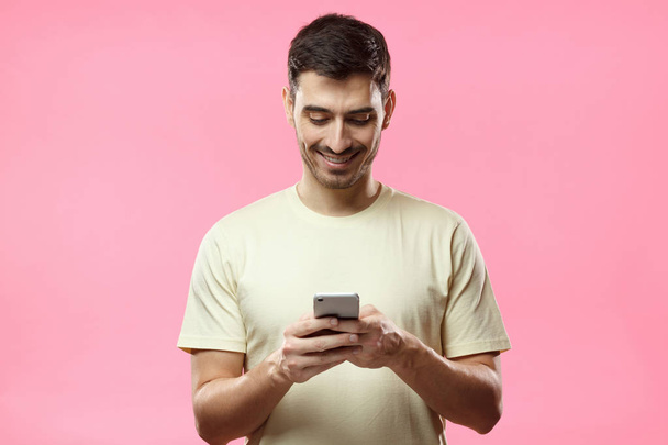 Indoor picture of young European Caucasian man pictured isolated on pink background smiling while looking attentively at screen of his smartphone, chatting or browsing with help of wireless connection - Zdjęcie, obraz