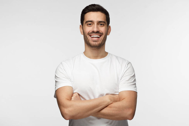 Half-length indoor portrait of young European Caucasian man isolated on gray background standing in white T-shirt with arms crossed, smiling and laughing positive as he is looking straight at camera - Φωτογραφία, εικόνα