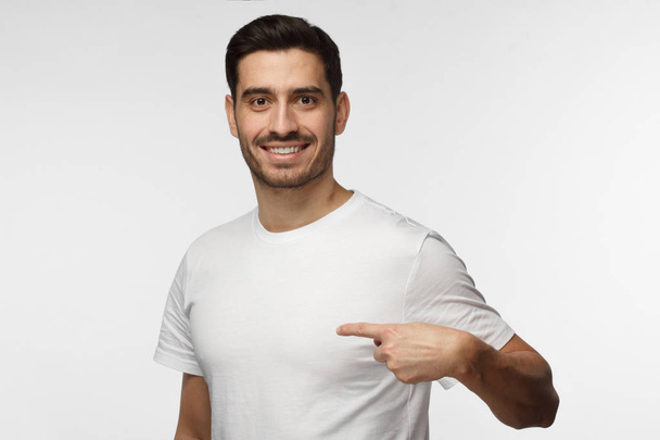 Horizontal picture of young handsome Caucasian man pictured isolated on grey background smiling positively while pointing with index finger to white blank T-shirt, copyspace for advertising - Photo, image