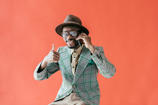 fashionable african american man with thumb up talking on smartphone, isolated on red  - Foto, imagen