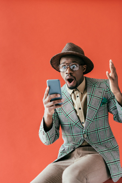 excited fashionable african american man using smartphone, isolated on red  - Фото, изображение