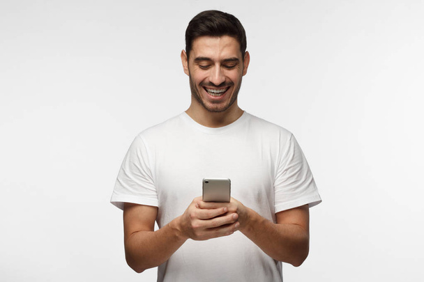Indoor picture of handsome Caucasian man isolated on gray background wearing white casual T-shirt laughing happily being amused by content on screen of smartphone he is holding in both hands - Photo, Image