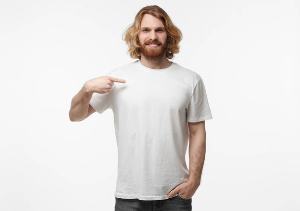 Half-length portrait of young handsome bearded male with long fair hair isolated on white background dressed in white blank T-shirt pointing to it with finger of right hand, copyspace for advertising - Φωτογραφία, εικόνα
