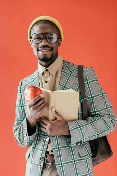 african american student in retro jacket with book and apple, isolated on red - Photo, Image