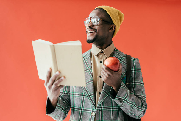stylish laughing african american man with book and apple, isolated on red - Fotoğraf, Görsel