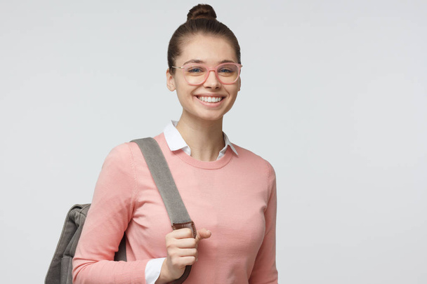 Closeup headshot of young beautiful European lady pictured isolated on grey background in eyeglasses showing positive smile while pulling forward strap of her grey backpack as if going to study - Fotó, kép