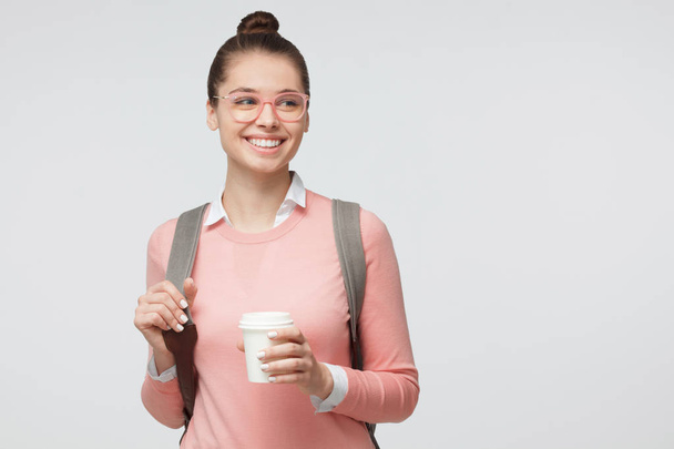 Photo d'intérieur de jeune femme caucasienne européenne isolée sur fond gris portant des lunettes et vêtue de façon décontractée et lumineuse avec un sac à dos derrière, tenant une tasse de café à emporter et souriant joyeusement
 - Photo, image
