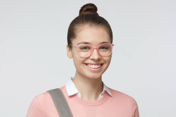 Indoor closeup of young pretty teenage girl pictured isolated on gray background wearing hipster pink eyeglasses, backpack strap crossing her shoulder, wearing hairbun, smiling friendly at camera - Foto, Imagem
