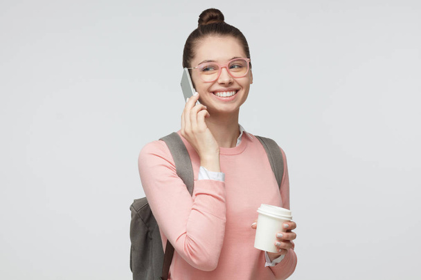 Closeup picture of young beautiful European lady isolated on grey background wearing casual clothes, pink trendy glasses and grey bag, feeling content and positive while having phone conversation - Foto, immagini