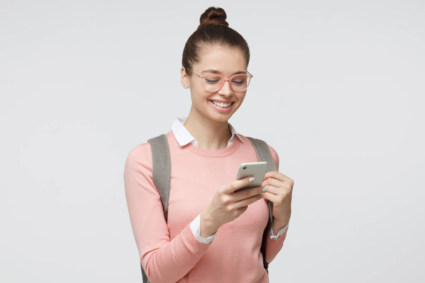 Indoor closeup of young European Caucasian girl pictured isolated on gray background with backpack on as if just returned from studies, looking at phone screen with nice smile, typing or browsing - Valokuva, kuva