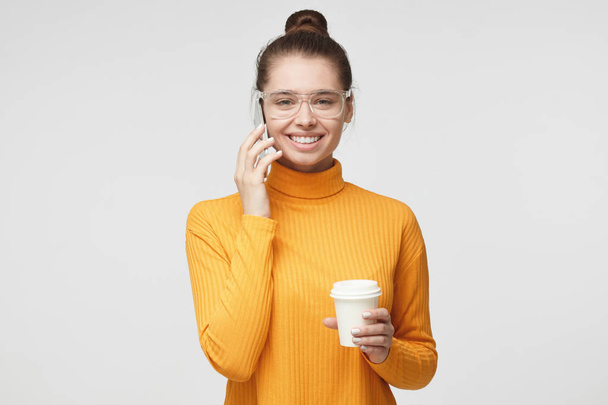 Closeup photo of young pretty European female isolated on gray background standing in bright yellow sweater with cardboard coffee cup, talking on cellphone with mate, showing happy friendly smile - Photo, Image