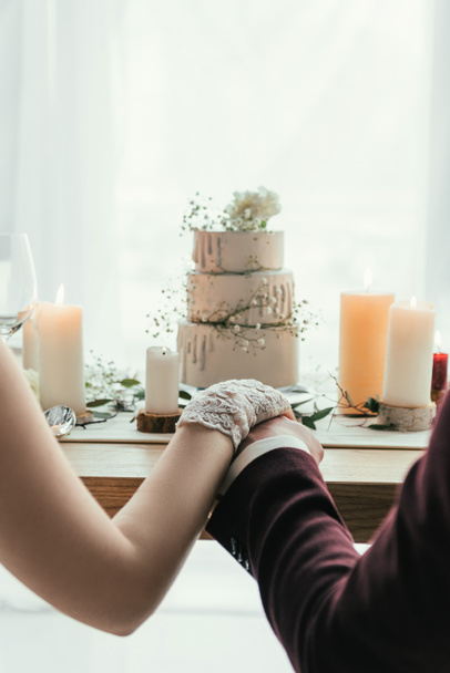 partial view of newlyweds holding hands while sitting at served table with wedding cake, rustic wedding concept - Photo, Image