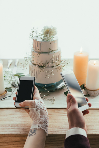 vista parcial de los recién casados usando teléfonos inteligentes con pantallas en blanco mientras están sentados en la mesa servida, concepto de boda rústico
 - Foto, Imagen