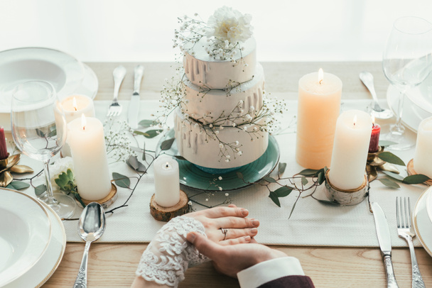 partial view of newlyweds holding hands while sitting at served table with wedding cake, rustic wedding concept - Photo, Image