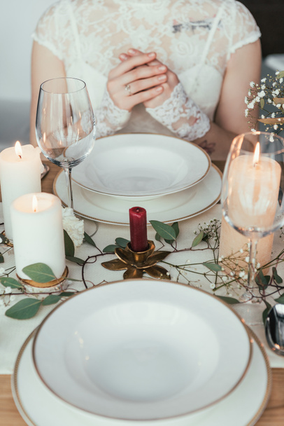 tiro recortado de novia en vestido blanco sentado en la mesa servida con platos vacíos y gafas de vino, concepto de boda rústico
 - Foto, Imagen