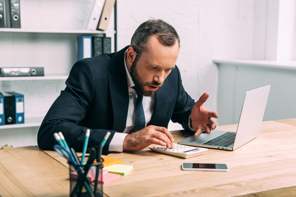 шокированный и напряженный бизнесмен с калькулятором на рабочем месте с ноутбуком в офисе
 - Фото, изображение