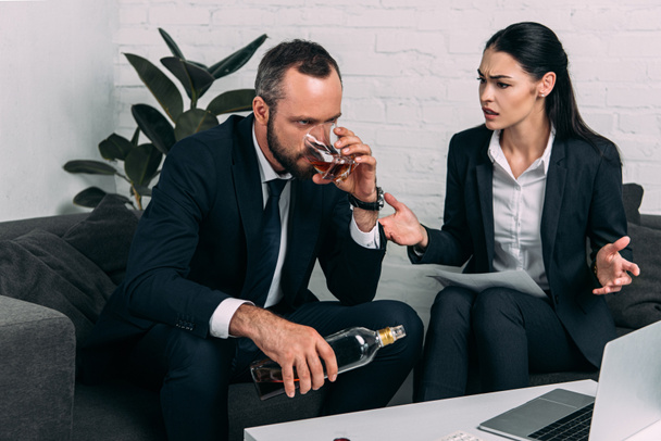 stressed businessman with alcohol sitting near colleagues at coffee table with laptop in office - Foto, immagini