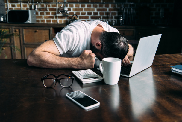 freelancer cansado trabajando a distancia tumbado en la mesa con el ordenador portátil, calculadora y taza de café en la cocina en casa
 - Foto, Imagen